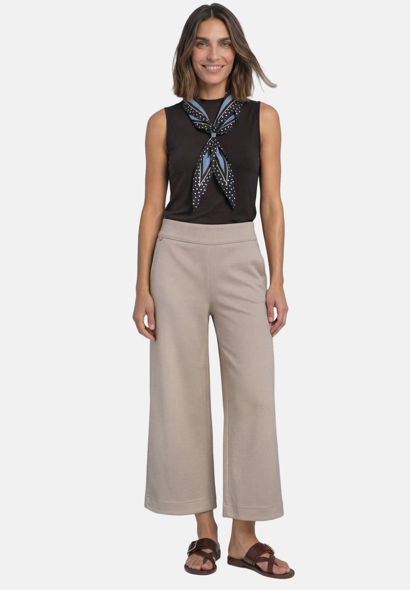 Femme portant un haut noir sans manches, un foulard à pois, un pantalon beige à jambes larges et des sandales marron, debout et souriante sur fond blanc.