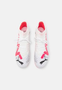Scarpe atletiche Puma bianche con accenti rosa. Presentano una tomaia texturizzata, un design elegante, lacci e un logo sulla punta.