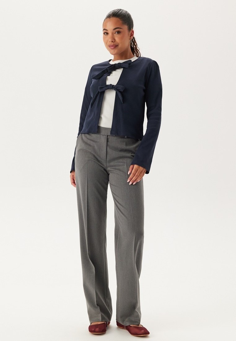 Marinefarbener verkürzter Cardigan mit Schleifendetail, getragen über einem weißen Oberteil, kombiniert mit grauen taillierten Hosen und kastanienbraunen Ballerinas. Glatte Textur; taillierte Passform.