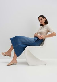 Jupe maxi en denim bleu avec une forme évasée, assortie à un haut crème et des sandales à talons beiges ; assise sur une chaise blanche incurvée.