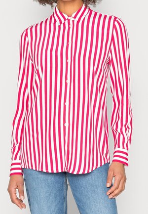 Femme portant une chemise à boutons à rayures verticales rouges et blanches à manches longues avec un jean bleu, debout devant un fond clair.