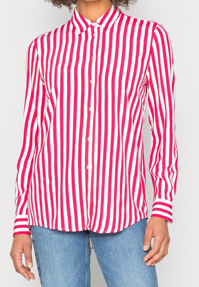 Femme portant une chemise à boutons à rayures verticales rouges et blanches à manches longues avec un jean bleu, debout devant un fond clair.