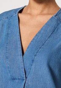 Gros plan d'une personne portant un vêtement en denim bleu à col en V, avec un col visible et des détails de coutures.