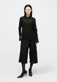 Camisa preta transparente de botão com mangas longas, combinada com calças pretas largas arregaçadas na bainha. Sapatos de salto pretos complementam o conjunto.