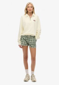 Sudadera con cremallera de color amarillo claro con un pequeño logo, combinada con pantalones cortos de cuadros verdes. El modelo lleva calcetines blancos y zapatillas beige.