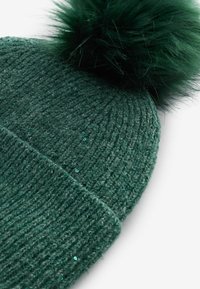 Kootud roheline müts tekstuurse mustriga, millel on pehme pom-pom üleval ja väikesed sädelevad detailid.