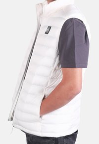 Personne portant un gilet matelassé blanc sans manches, les mains dans les poches, sur une chemise sombre à manches courtes, sur un fond uni.