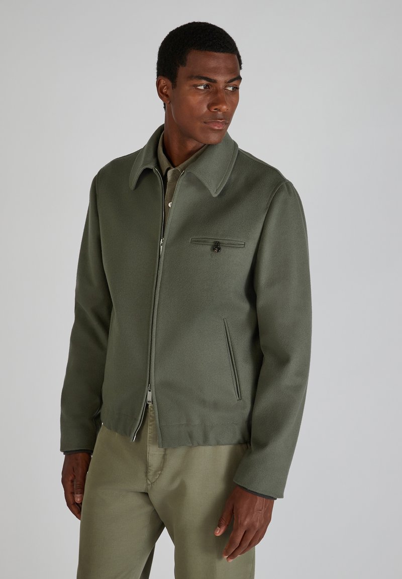 Olivegrüne Wollmischungjacke mit Reißverschluss vorne, weit ausgestelltem Kragen und einer Brusttasche. Glatte Textur und minimale Hardware-Akzente.