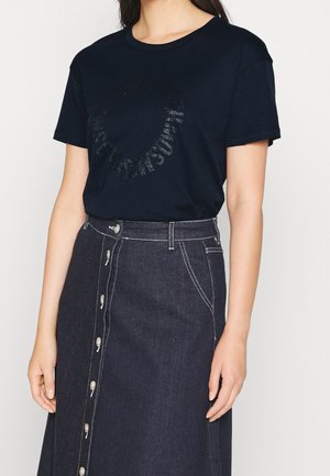 Femme portant un t-shirt noir à manches courtes avec un texte circulaire subtil et une jupe en jean foncé taille haute à boutons sur le devant.