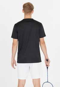 Svart atletisk t-shirt med korta ärmar, enkel design. Bärs med vita shorts; person håller en badmintonracket i höger hand.