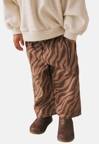 Dziecko ubranie w beżowy sweter w kropki, brązowe spodnie w zebry i błyszczące brązowe botki, stojące z jedną ręką w kieszeni.