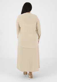 Abito beige a maniche lunghe con orlo arrotondato e una superficie liscia, caratterizzato da una leggera silhouette ad A. Tacchi a stiletto visibili sotto.