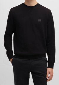 Sweat-shirt noir en matière douce, avec un col rond, des poignets côtelés et une petite étiquette de marque sur la poitrine.