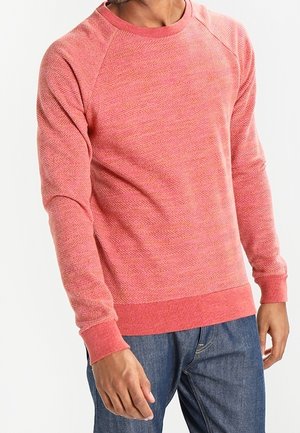 Mężczyzna ubrany w koralowo-różowy sweter z długim rękawem o fakturowanej powierzchni oraz niebieskie dżinsy, stojący ze swobodnie opuszczonymi ramionami.