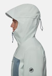 Veste imperméable gris clair avec une grande capuche, ornée d'une accent bleu à l'avant, fermeture éclair, et un patch logo sur la manche.