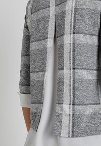 Veste à carreaux grise avec une coupe décontractée, dotée d'une découpe fendue dans le dos et de manches trois-quarts. Tissu texturé avec des accents blancs.