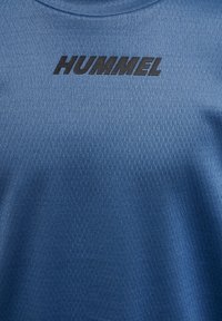 Blauer strukturierter Stoff mit dem "HUMMEL"-Logo, das in fetten schwarzen Buchstaben über der oberen Mitte aufgedruckt ist.