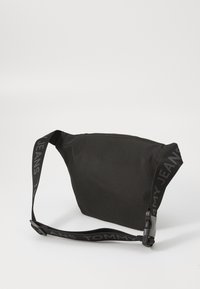 Schwarze Stoff Schultertasche mit verstellbarem Tragegurt, die das "TOMMY JEANS"-Logo zeigt, minimalistisches Design und glatte Textur.