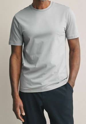 Maglietta grigia a maniche corte con scollo rotondo, realizzata in tessuto morbido, caratterizzata da una texture liscia e un design aderente, abbinata a pantaloni scuri.