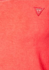 Koszula w kolorze koralowym o gładkiej fakturze. Posiada małe trójkątne logo "GUESS" w lewym górnym rogu. Prosty design i jednolity kolor.