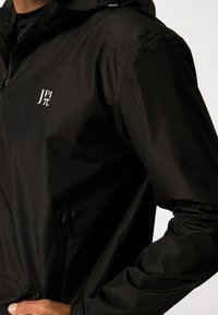 Zwarte jas van waterafstotend materiaal, met een ritszak, capuchon en een wit logo op de borst. Soepele textuur, minimalistisch ontwerp.