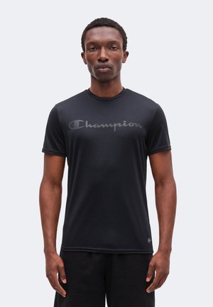 Mann steht frontal, trägt ein schwarzes Champion-T-Shirt mit Logo auf der Brust und schwarze Shorts vor einem einfarbigen hellen Hintergrund.