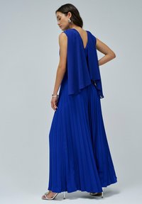 Vestido de noche largo y plisado en tela azul royal con un detalle de capa fluida en los hombros y un diseño de espalda descubierta. Accesorios plateados visibles.