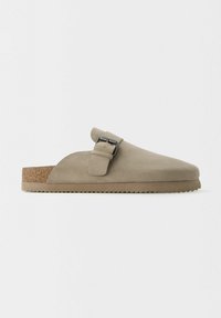 Chaussure en suède beige à enfiler avec une semelle en liège, bout rond et une sangle sur le côté avec un accent en boucle métallique. Dessus texturé et semelle crantée.