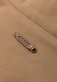 Gros plan d’un tissu beige avec une couture cousue et une étiquette en cuir marron attachée indiquant « ombre QUIETESSENCE. »