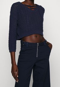 Marinblå kort stickad tröja med ett öppen vävmönster och detaljerad halsringning, i kombination med högmidjade marinblå byxor som har dragkedja och bälteshällor.