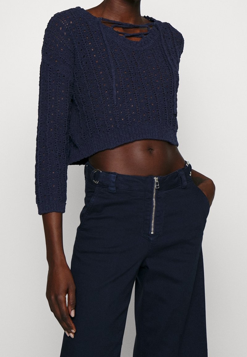 Marinblå kort stickad tröja med ett öppen vävmönster och detaljerad halsringning, i kombination med högmidjade marinblå byxor som har dragkedja och bälteshällor.
