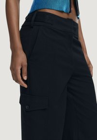 Close-up van een persoon in zwarte cargobroek met zijflapzakken en een blauwe crop top die de buik laat zien, met een ontspannen hand.