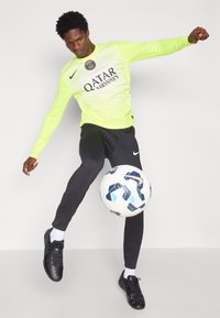 Nike neona dzeltena garā piedurkne kreklā ar horizontālām svītrām, melni bikses, melnā futbolā, un rakstains futbola bumba.