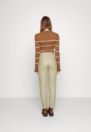 Vrouw die met haar rug naar de camera staat, draagt een bruin gestreept topje met lange mouwen, lichtbeige broek en bruine stevige schoenen tegen een effen achtergrond.