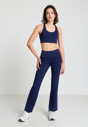 Frau steht in einem marineblauen Sport-BH und passenden weiten Leggings mit weißen Turnschuhen vor einem einfarbigen grauen Hintergrund.