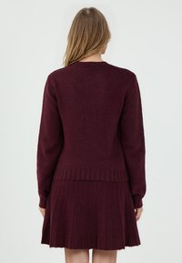 Burgunderpullover mit geripptem Saum, langen Ärmeln und rundem Halsausschnitt, kombiniert mit einem plissierten Rock in passender Farbe. Weiche Textur.