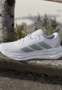 Chaussure de sport blanche avec des rayures vert clair posée sur un rebord en pierre à l'extérieur, avec des plantes floues et un tronc d'arbre en arrière-plan.