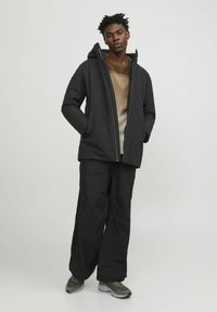 Chaqueta aislante negra con capucha, presenta un exterior liso, bolsillos laterales y un cierre frontal. Combinada con pantalones negros holgados y zapatillas.