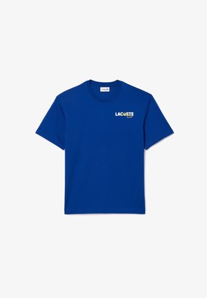 T-shirt bleu roi avec logo Lacoste, pantalon beige, sac à bandoulière beige et baskets blanches avec accents bleu clair. Fond uni.