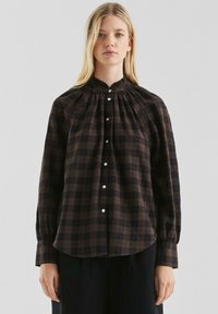 Bruine geruite blouse met hoge hals, geplooide details en knoopsluiting. Lange mouwen met knoopmanchetten, gemaakt van een lichtgewicht stof.