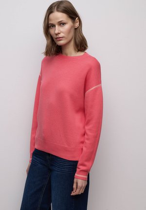 Donna con capelli castani fino alle spalle che indossa un maglione a maniche lunghe rosa corallo e jeans blu scuro, in piedi davanti a uno sfondo grigio chiaro.