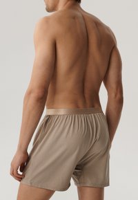 Beżowe bawełniane bokserki z elastycznym pasem, z gładką fakturą i luźnym krojem. Model widziany z tyłu.