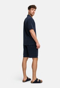 Tiefblaues Poloshirt und Shorts-Set aus Frottee mit lockerer Passform. Merkmale sind kurze Ärmel, schlichtes Design und passende Schlippersandalen.