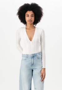 Bílý bodysuit s dlouhým rukávem má hluboký výstřih do V, texturovaný krajkový vzor a je zkombinovaný s vysokými modro-světle modrými džínami.