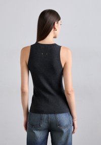 Kvinna med långt brunt hår som bär en ärmlös mörkgrå topp och blå jeans, står vänd mot en enkel ljusgrå bakgrund.