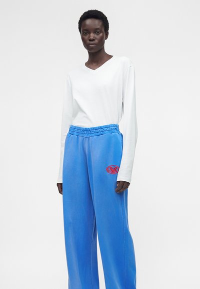 Άτομο που φοράει λευκό μακρυμάνικο μπλουζάκι με V λαιμόκοψη και φαρδιά μπλε παντελόνια με κόκκινο κεντητό έμβλημα στο μηρό, στέκεται μπροστά σε απλό φόντο.