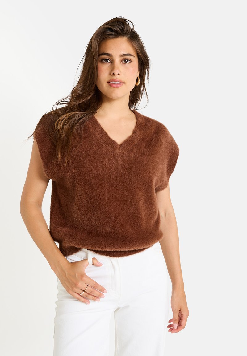 Pull en maille marron et duveteux avec un col en V et des manches courtes, associé à un jean blanc. Texture douce et coupe ample, détails de design minimalistes.