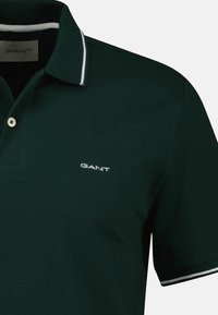 Ciemnozielona koszulka polo wykonana z bawełny, z kołnierzykiem wykończonym na biało, z trzema guzikami i małym białym logo na piersi.