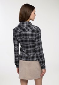 Camicia a quadri nera e grigia con maniche lunghe, polsini con bottone e una yoke nella parte posteriore. Il materiale sembra morbido e testurizzato, abbinato a una gonna beige.