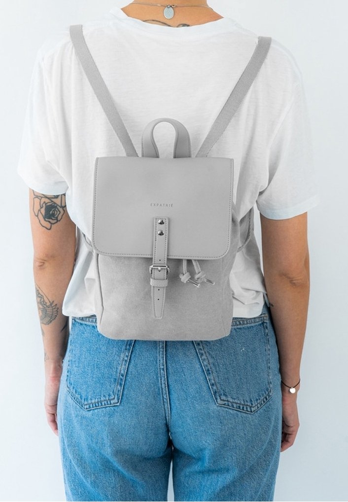 Personne portant un petit sac à dos gris clair par-dessus un t-shirt blanc et un jean bleu, montrant des tatouages sur le bras gauche et un collier.